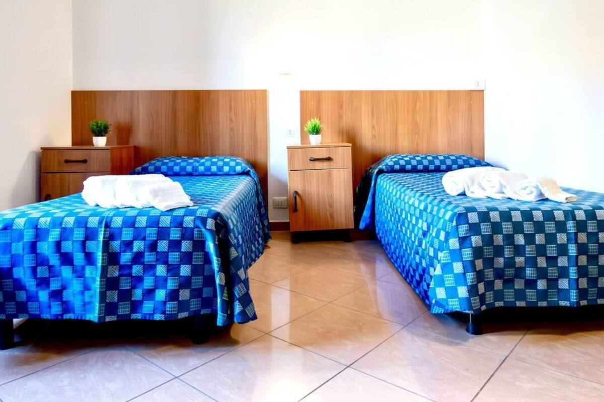 Habitación de hotel con dos camas, cada una con cubrecama azul a cuadros y toallas blancas. Poseen cabeceros de madera y una pequeña planta en cada mesita de noche.
