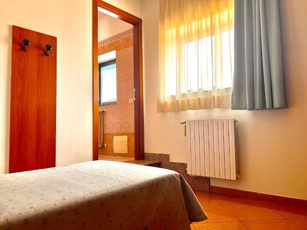 Habitación de hotel con cama, puerta de madera, ventana con cortinas, radiador y baño a la vista a través de una puerta abierta.