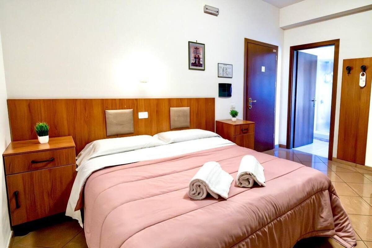 Una habitación de hotel con cabecera de madera, cama con ropa de cama rosa, dos mesitas de noche con una planta y una lámpara, un pequeño tocador y una puerta que da a un pasillo.