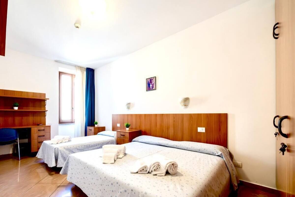 Habitación de hotel con dos camas, un escritorio, una silla y un pequeño ventanal con cortinas.