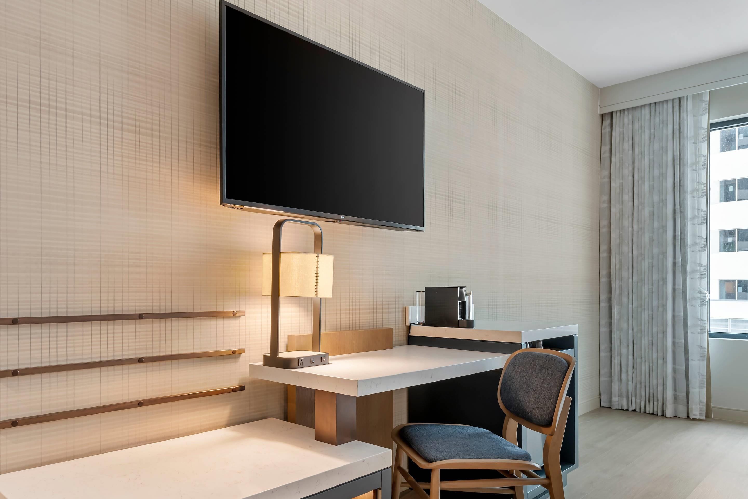 A hotel room with a large flat-screen TV mounted on the wall, a desk with a lamp, a chair, and a view of the city through a window with curtains.