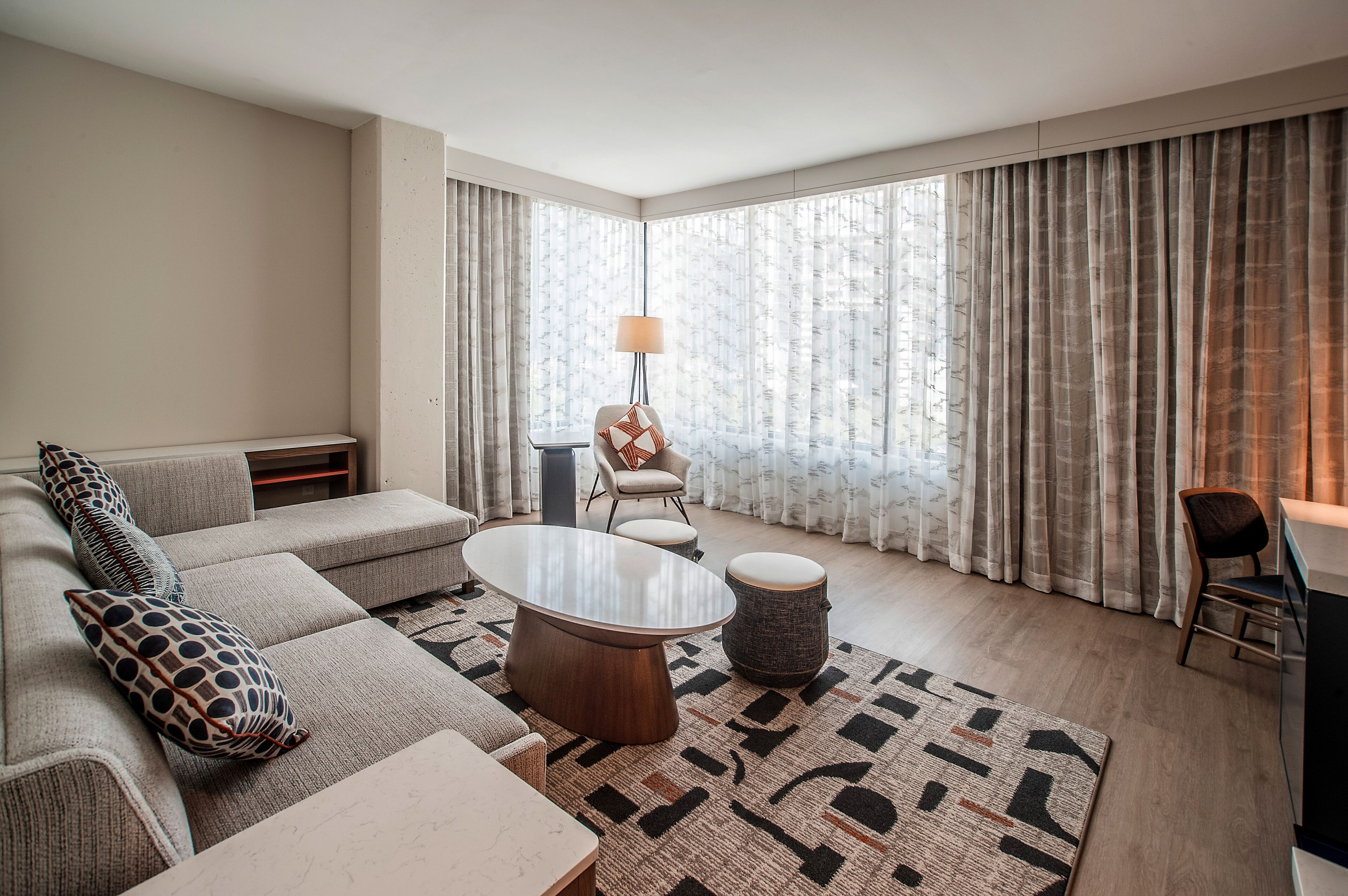Una sala de estar moderna con un sofá, una mesita de centro, una lámpara y vistas a través de ventanas grandes con cortinas.
