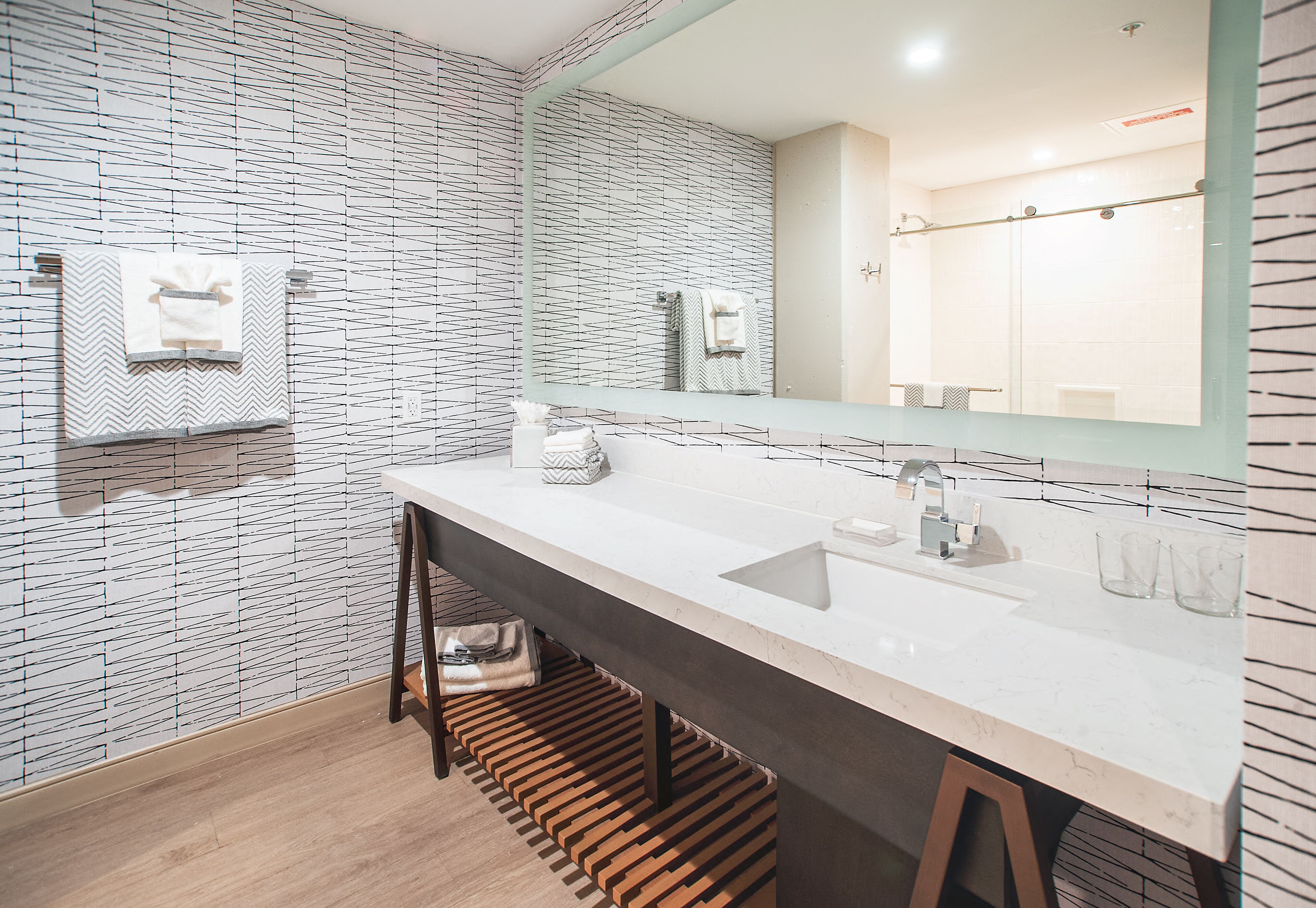 A modern bathroom with a white and black patterned wall, a large mirror, a white sink, and a wooden bench.