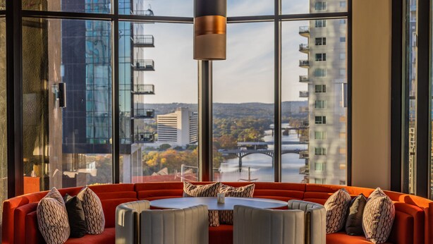 A modern interior with a red sofa, a round table, and a view of a cityscape through large windows. - Cambria Hotel Austin Downtown (Austin)