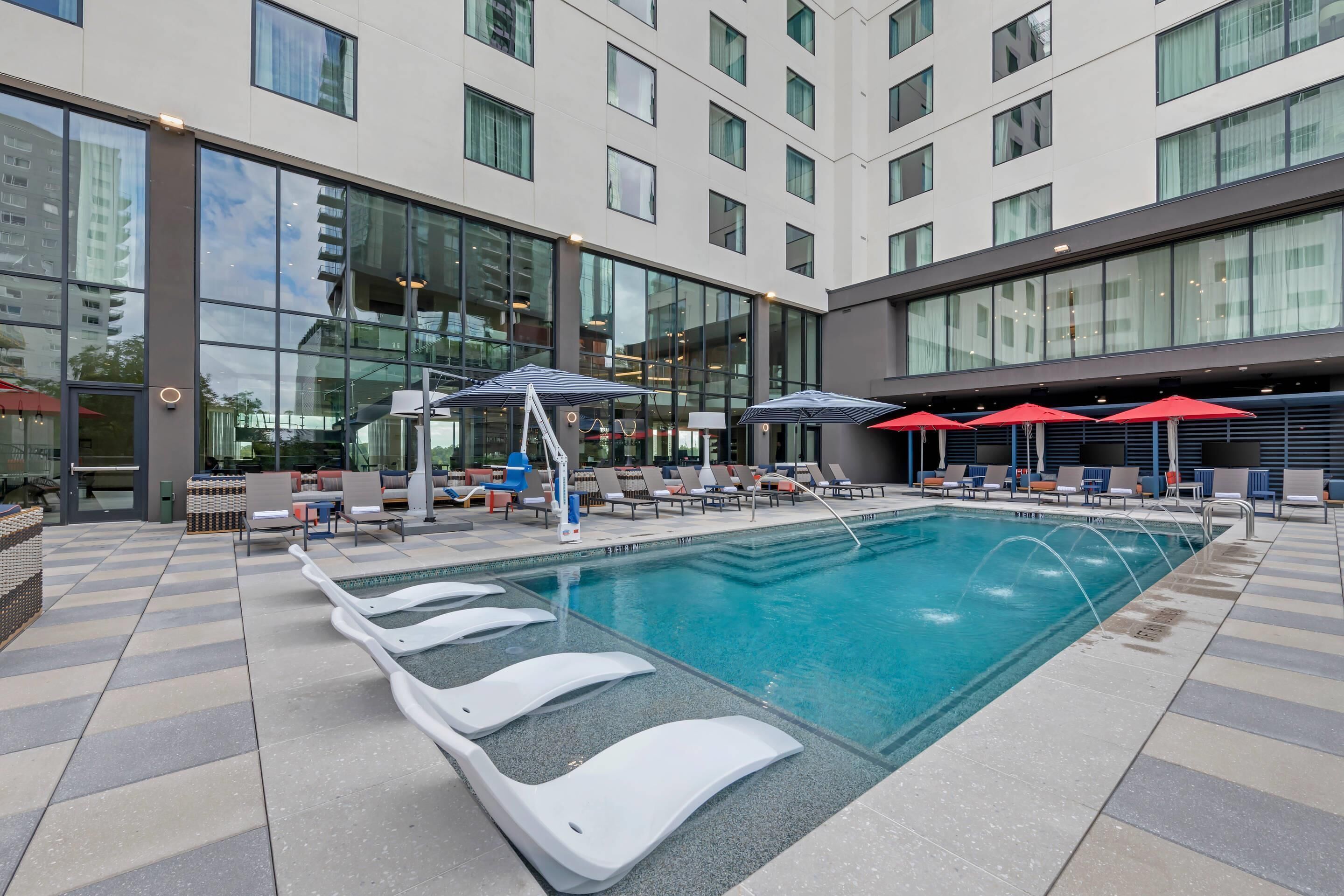 A modern hotel pool area with lounge chairs and umbrellas, large windows, and a clear blue pool.