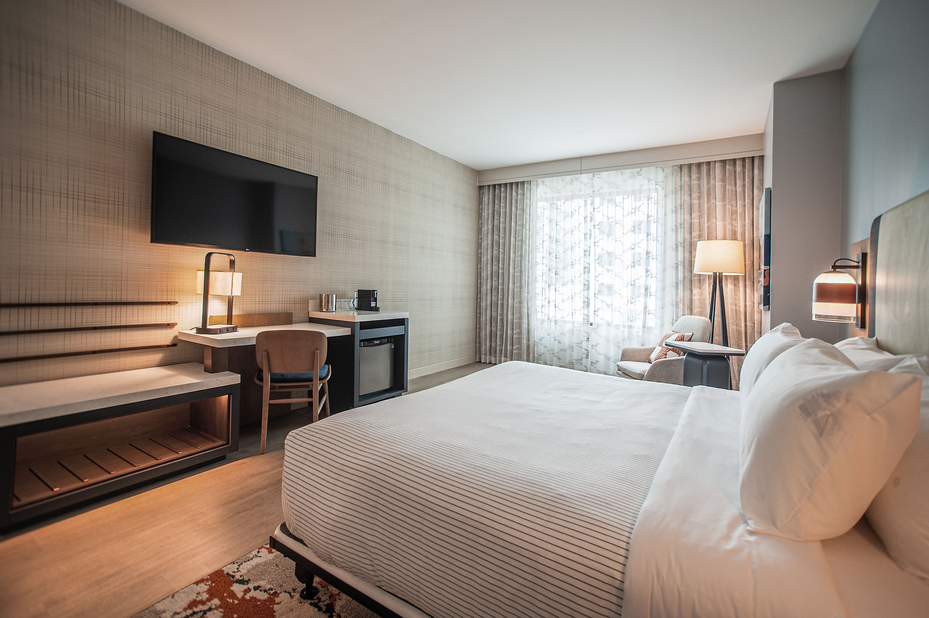 Une chambre d’hôtel avec un grand lit, un bureau avec une chaise, un téléviseur fixé au mur, une lampe et une fenêtre avec des rideaux.