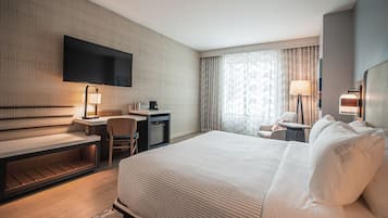 A hotel room with a large bed, a desk with a chair, a TV mounted on the wall, a lamp, and a window with curtains.