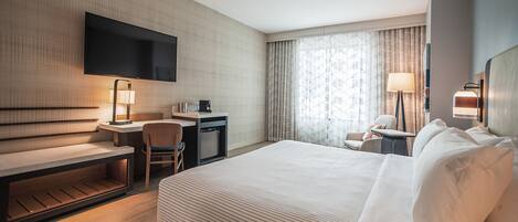 Une chambre d’hôtel avec un grand lit, un bureau avec une chaise, un téléviseur fixé au mur, une lampe et une fenêtre avec des rideaux.