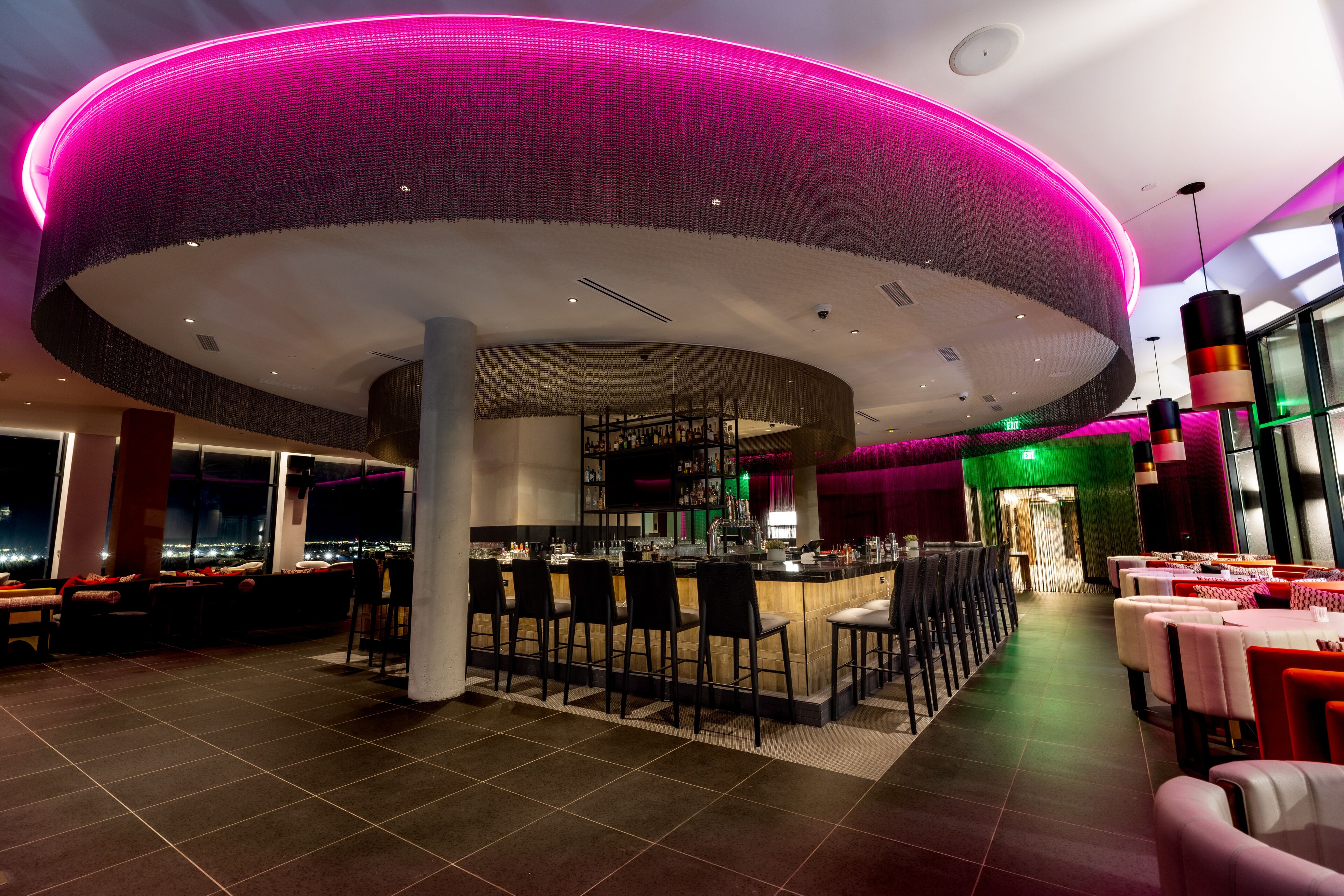 Een modern restaurant met een centraal bargedeelte, zitplaatsen en een uitzicht op de stadsgezicht door grote ramen.
