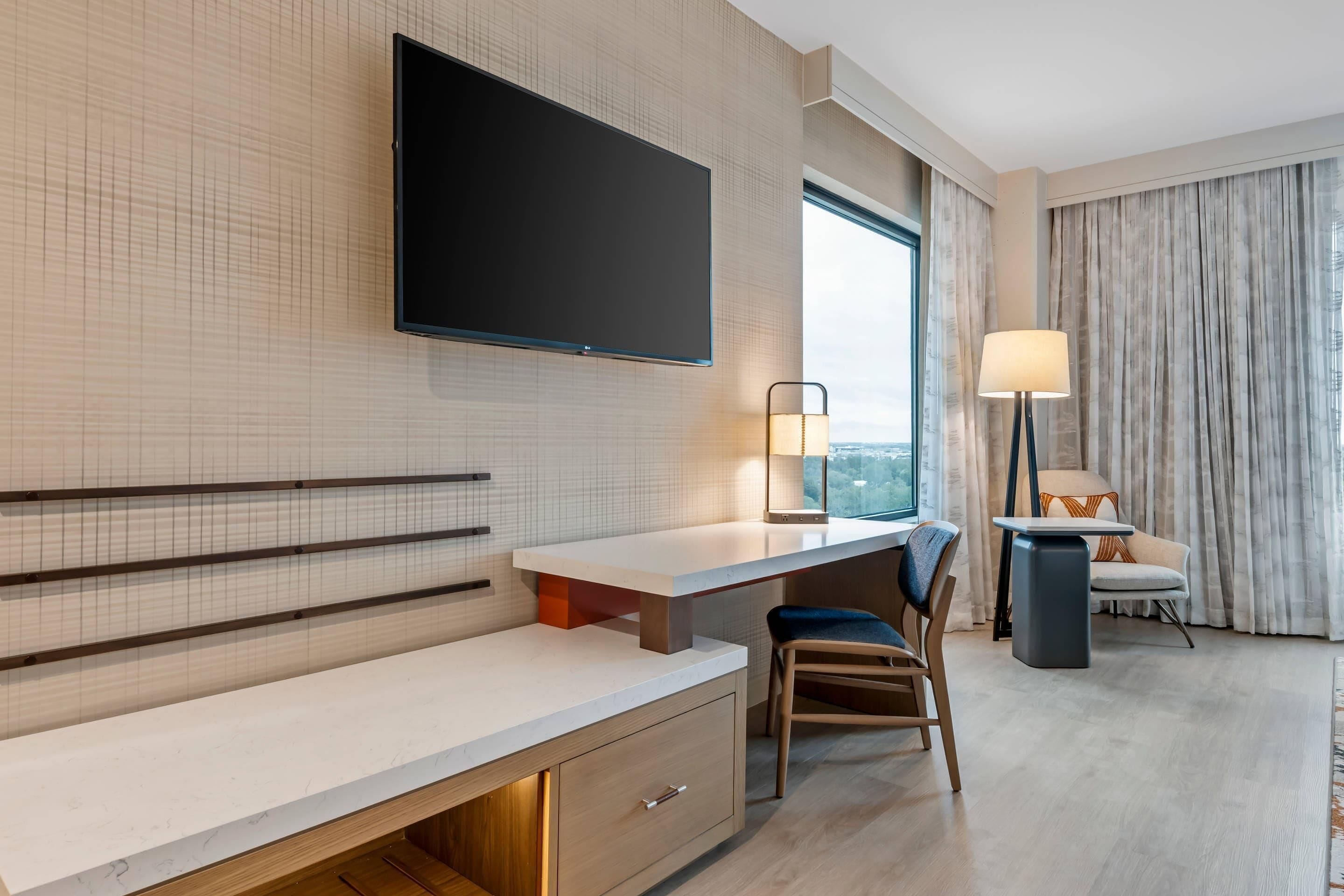 A hotel room with a flat-screen TV mounted on the wall, a desk with a chair, a lamp, and a view of the outdoors through a window.