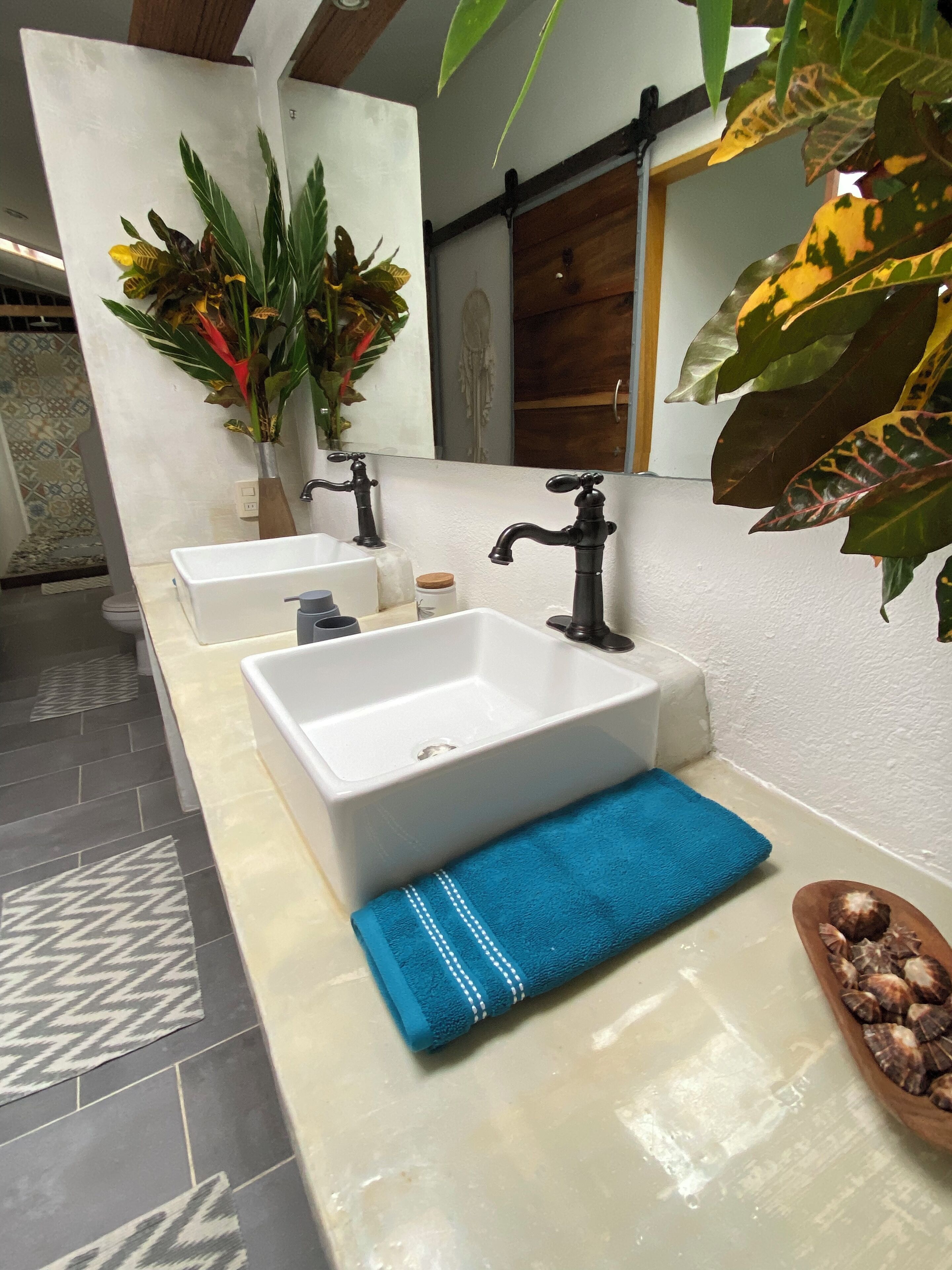 Un baño con dos lavamanos blancos, una toalla azul y un cuenco de madera con conchas marinas.
