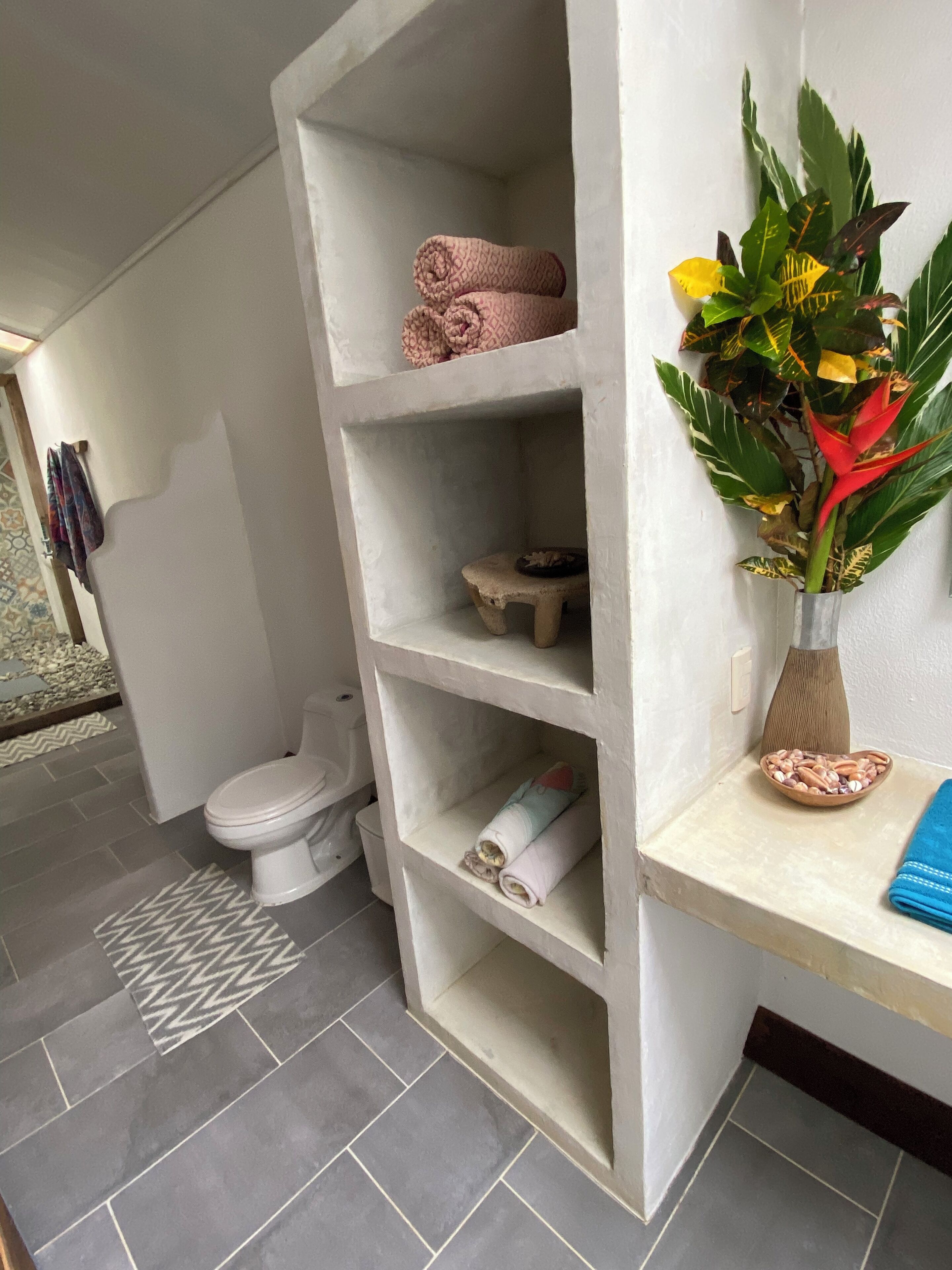 Et badeværelse med et hvidt toilet, et håndklædestativ, en hylde med håndklæder, en lille skammel og en vase med en plante.