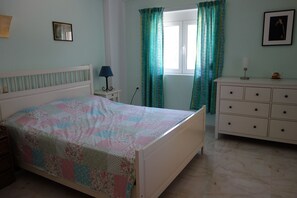 Une chambre à coucher comprenant un lit, une commode, une table de chevet, une lampe, une fenêtre avec des rideaux et un tableau encadré accroché au mur.
