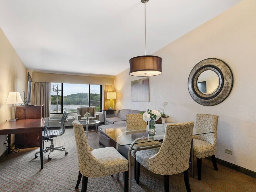 A hotel room with a desk, chairs, a sofa, a coffee table, and a large window offering a view.