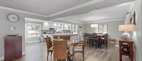 A spacious living area with a dining table and chairs, a kitchen in the background, and a clock on the wall.