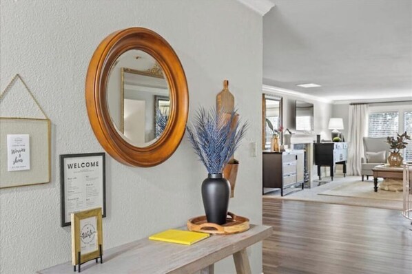 Un área de recepción con un banco de madera, un letrero de bienvenida, un espejo y un jarrón con ramas decorativas azules.