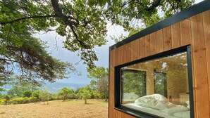 A modern wooden cabin with a large window, a bed inside, and a view of a grassy landscape with trees. - La Bastide Provençale - Terre D'ocre & de Lavande (Viens)