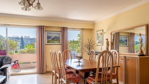 A dining area with a wooden table and chairs, large windows with curtains, and a view of buildings and greenery outside. - Flat with swimming pool close to the sea (Cagnes-sur-Mer)