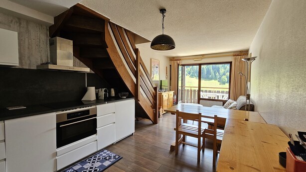 A modern kitchen with a wooden staircase, a dining area with a wooden table and chairs, and a large window offering a view of the outdoors.