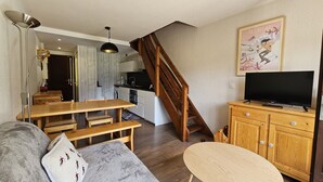 A cozy living room with a wooden staircase, a dining area, a kitchen, and a television.