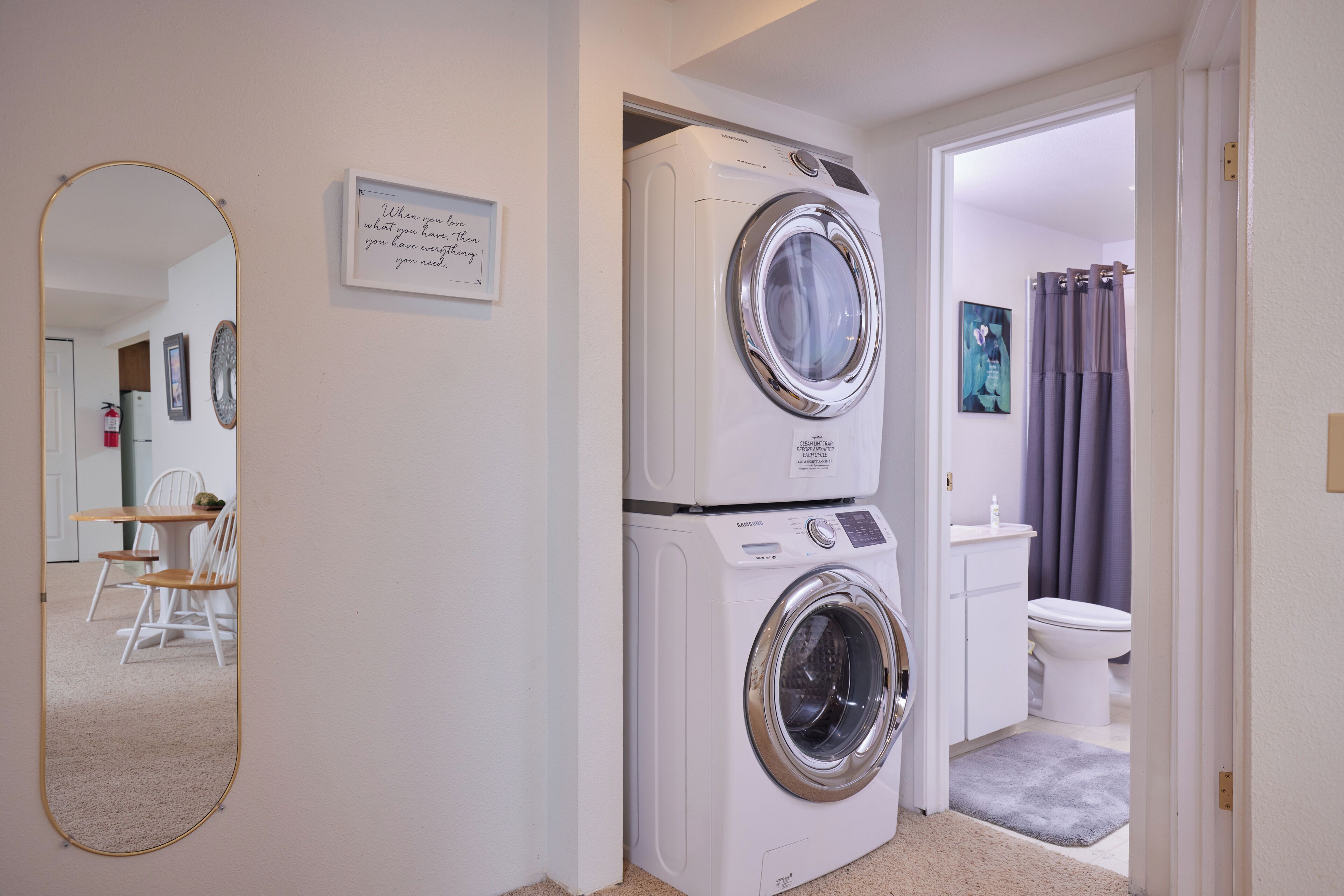 A laundry room with two stacked washers and dryers, a mirror, a framed inspirational quote, and a view into the bathroom.