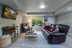 A living room with a leather sofa, a wooden coffee table, a chair, a TV mounted on the wall, and a window with a view of greenery.