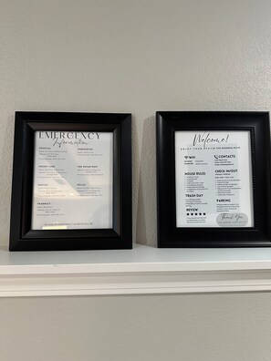 Two framed documents on a shelf: one titled 'EMERGENCY Information' listing various contacts and services, and the other titled 'Welcome!' with house rules and amenities.