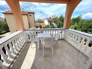 A balcony with a table and chairs, surrounded by a white balustrade, overlooking a residential area with houses and greenery. - Holiday apartment with balcony and air conditioning (Jadranovo)