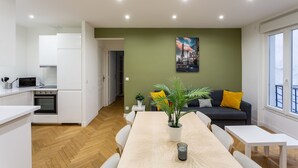 Eine moderne Wohnung mit Küche, Essbereich und Wohnzimmer. Sie verfügt über ein großes Fenster, ein Stadtbildgemälde und einen Blumentopf.
