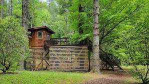 A cabin with a wooden exterior, a small window, and a staircase leading up to a deck. - Riverbright Treehouse on the Toccoa (Suches)