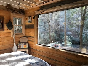 A rustic cabin room with a large window, wooden walls, a bed, a chair, and a vase. - Riverbright Treehouse on the Toccoa (Suches)