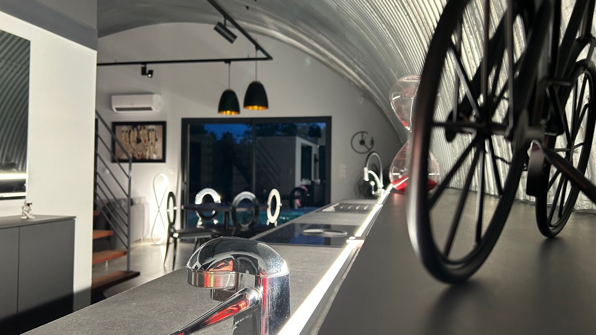 A modern kitchen with a long countertop, a bicycle wheel in the foreground, and a staircase leading to an upper level.