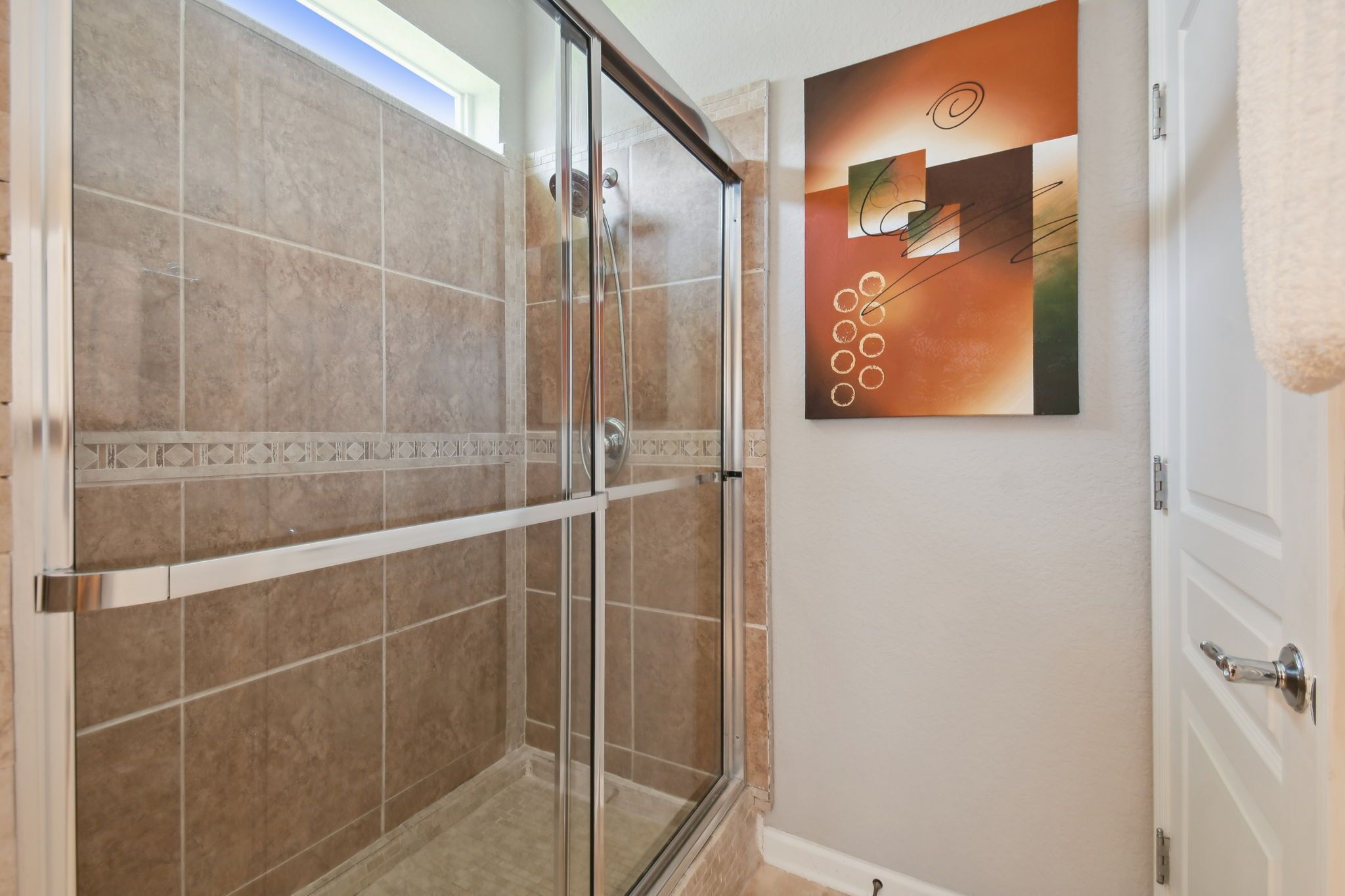 Ducha con puertas de vidrio y azulejos color beige, puerta blanca con una toalla colgada, y pared con un cuadro abstracto geométrico.