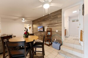 Un coin repas avec une table en bois, des chaises, un ventilateur de plafond et un téléviseur fixé au mur.