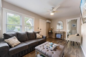 A living room with a grey sofa, a matching ottoman, a ceiling fan, and a door leading to another room.
