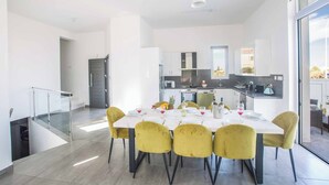 A modern dining area with a white table set for six, yellow chairs, and a kitchen in the background. - Villa Polymnia Protaras 1 (Paralimni)