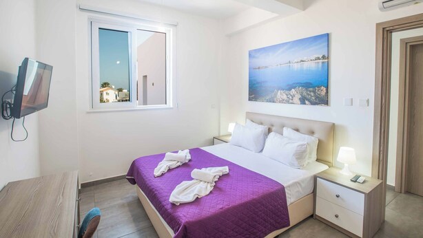 A hotel room with a bed, a nightstand with a lamp, a TV mounted on the wall, and a window with a view of a building. - Villa Polymnia Protaras 1 (Paralimni)