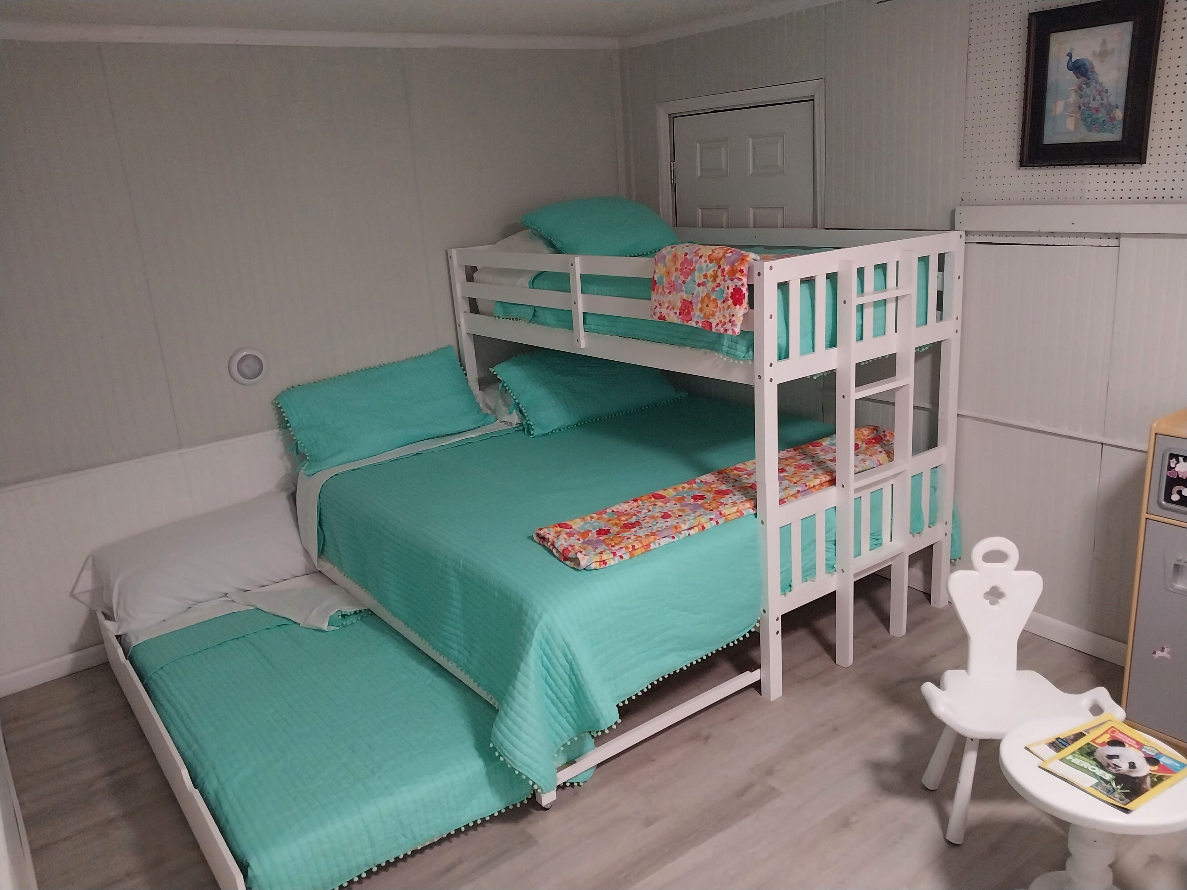 Habitación con literas, ropa de cama color turquesa, una puerta blanca, un cuadro enmarcado y una mesita con una revista.