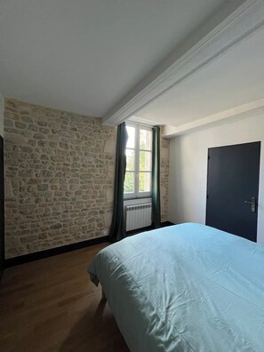 A bedroom with a stone wall, a window with curtains, a radiator, and a bed with a blue bedspread.
