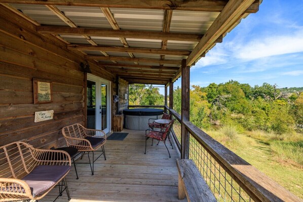 Una cabaña de madera con un porche cubierto, sillas de mimbre, un jacuzzi y vistas del bosque circundante.