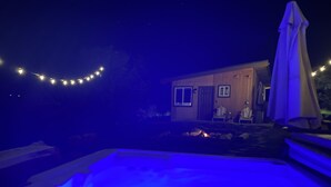 Un área de piscina con un elemento acuático iluminado en azul, una zona de estar cubierta y un edificio con puerta y ventanas.