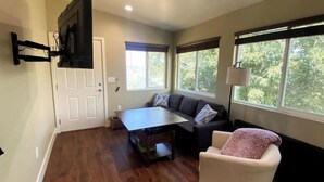 Una sala de estar con un sofá, una silla, una mesa de centro, un televisor montado en la pared y ventanas grandes con vistas a árboles.
