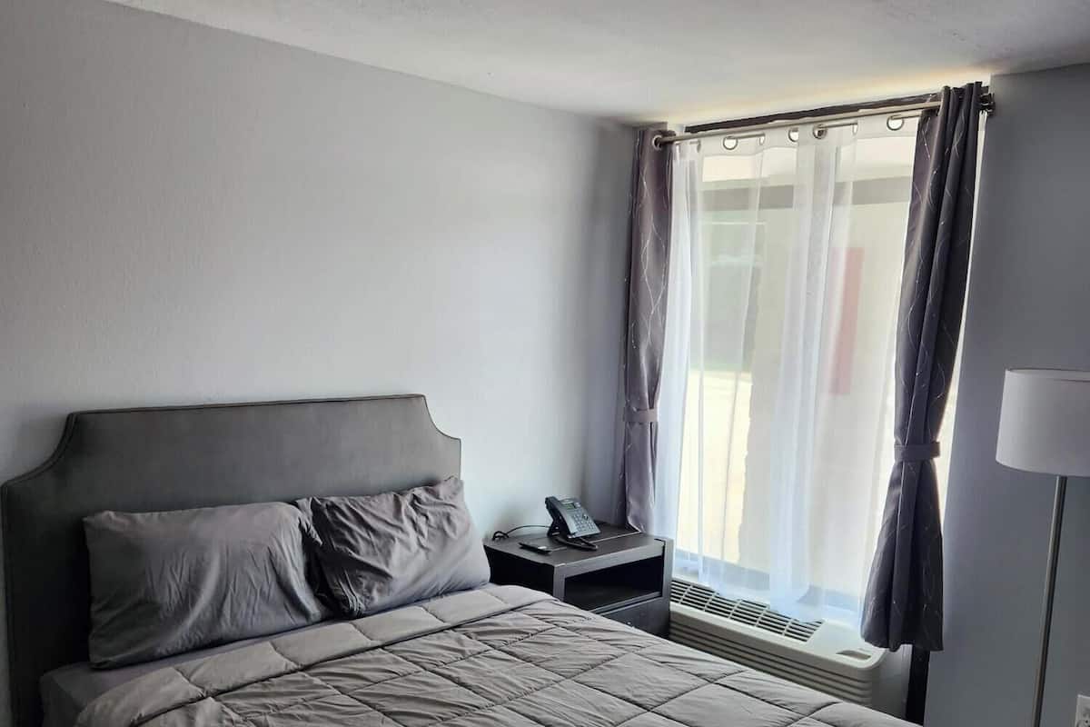 Een hotelkamer met een bed, nachtkastje met telefoon, raam met gordijnen en een radiator.