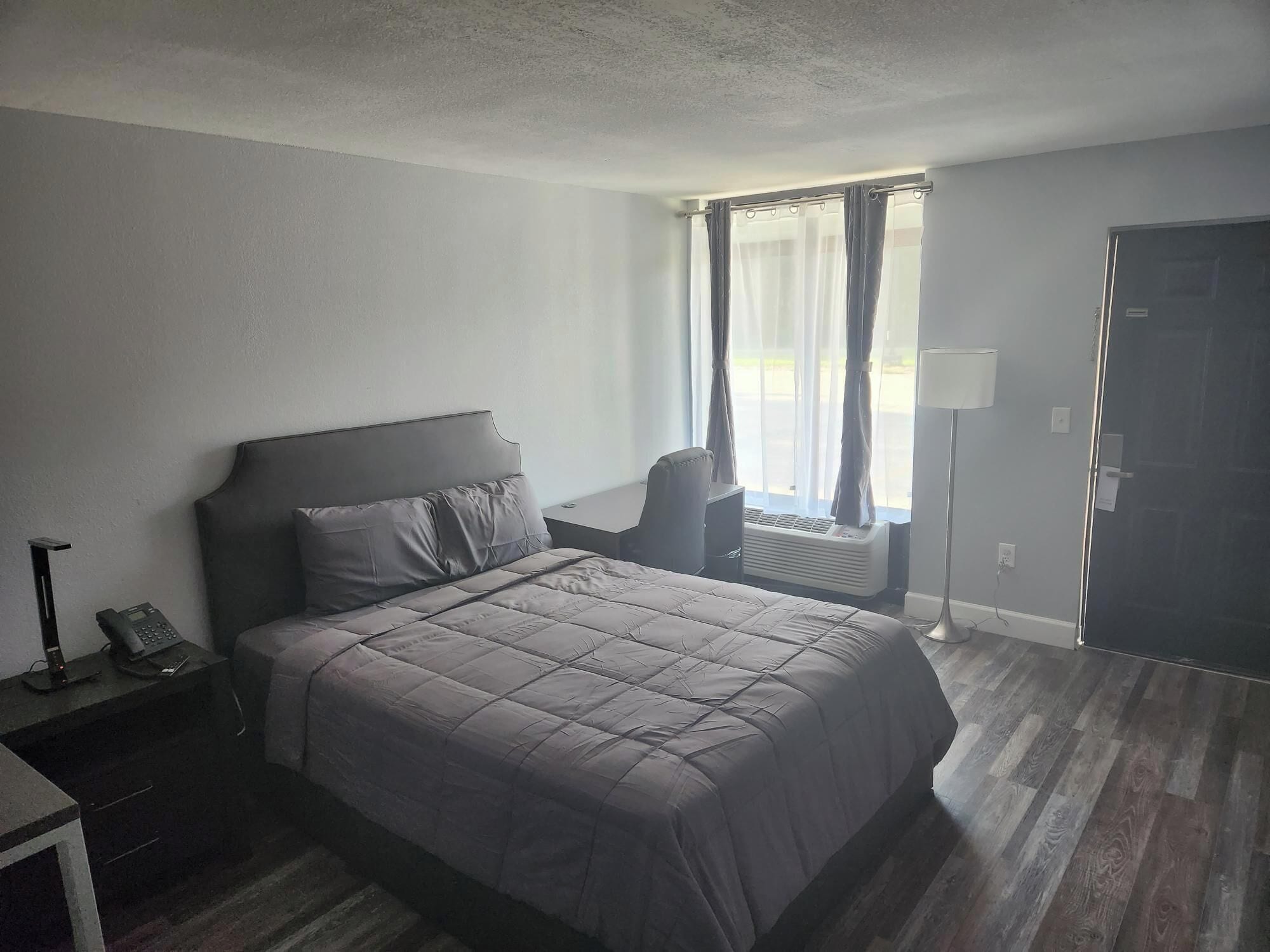 A hotel room with a bed, a desk with a phone, a chair, a window with curtains, and a door.