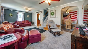 Ein Wohnzimmer mit einem roten Ledersofa, einem roten Hocker, einem Couchtisch aus Holz und einem Deckenventilator. Eine Treppe führt zu einer weiteren Ebene, und eine amerikanische Flagge ist an der Wand sichtbar.