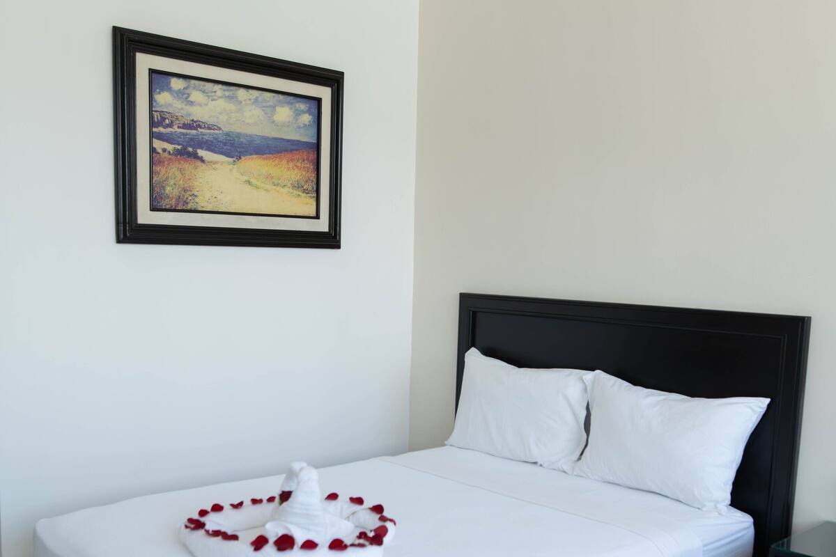 A neatly made bed with white linens and a decorative rose arrangement, a framed painting on the wall, and a black nightstand beside the bed.