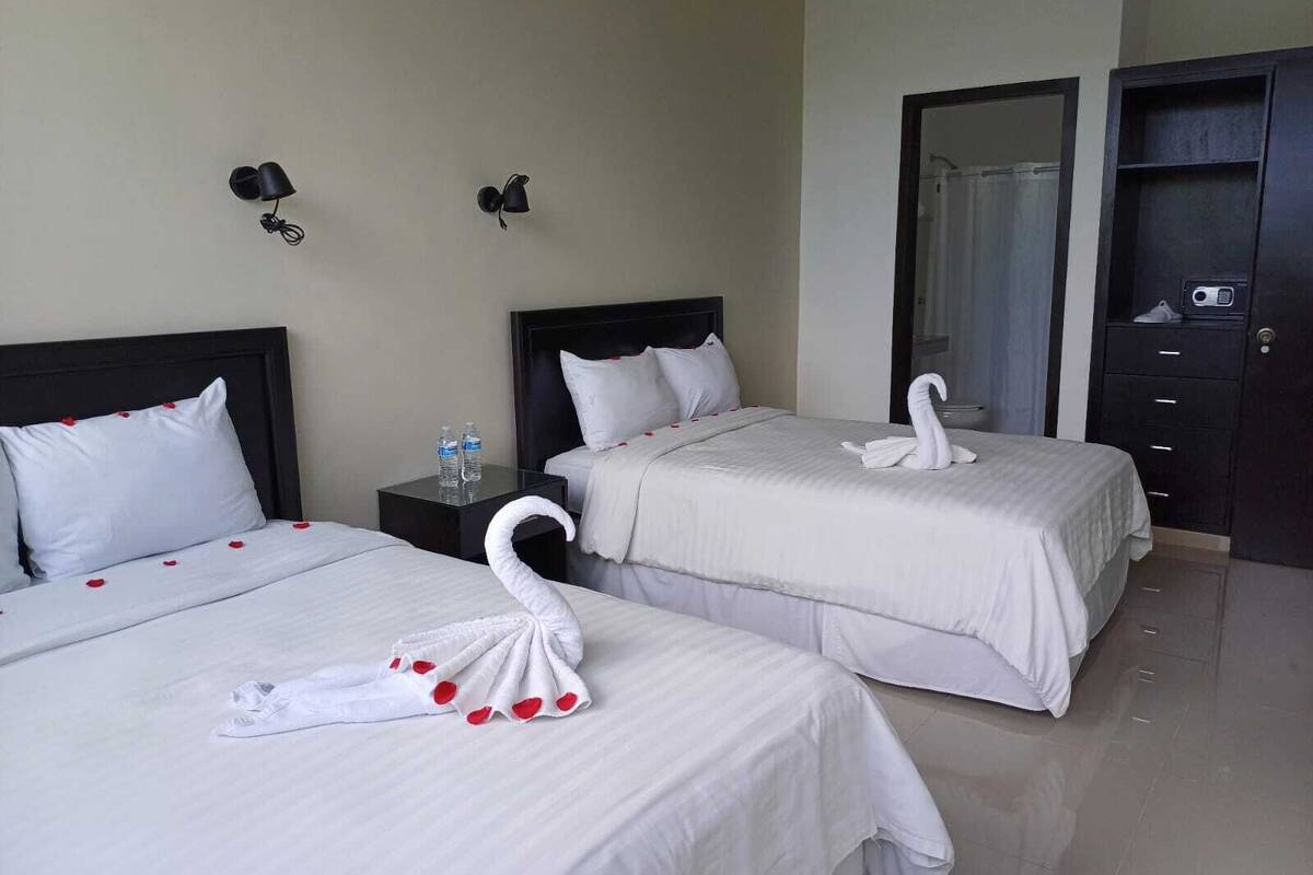 A hotel room with two beds, each with white bedding and red rose petals. There are two wall-mounted lamps and a dark-colored wardrobe.