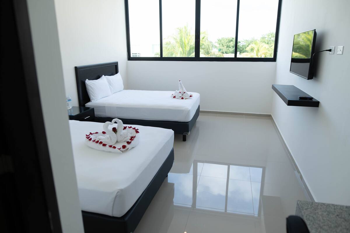 A hotel room with two beds, a TV mounted on the wall, and a window with a view of greenery.