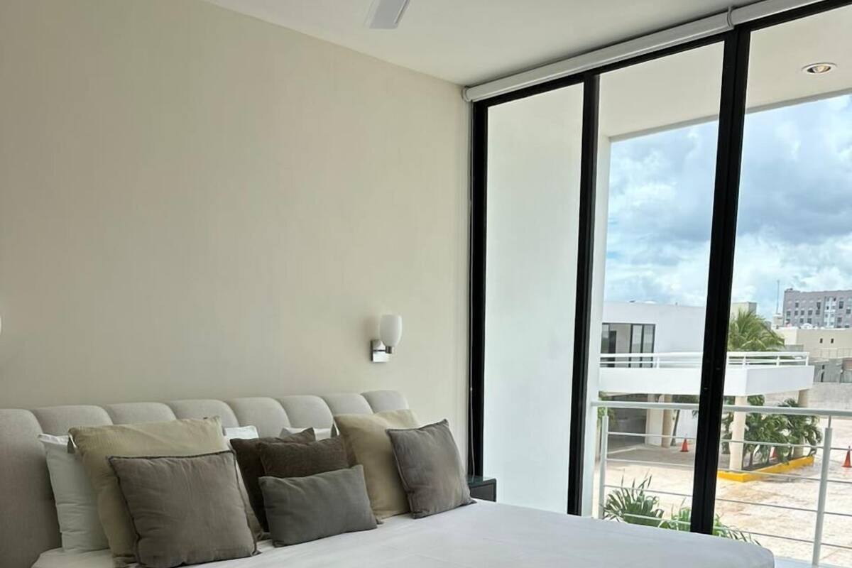A modern bedroom with a large bed, a wall-mounted light, and a view of the city through large windows.