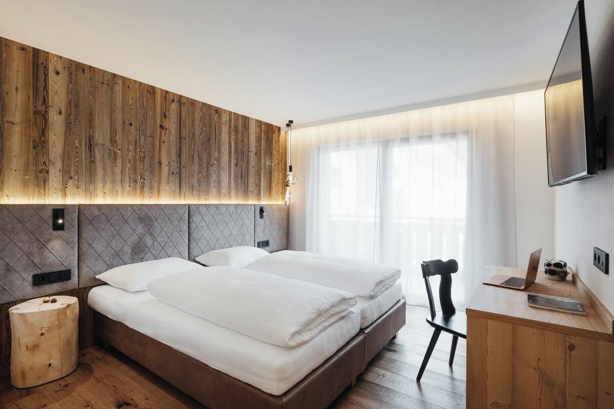 Quarto de hotel moderno com cabeceira de madeira, uma cama grande, uma escrivaninha com um laptop e uma TV na parede.